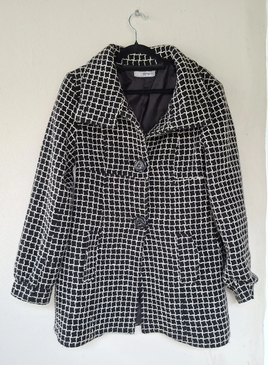 Abrigo de paño con un estampado cuadriculado en blanco y negro. Posee cuello solapa, dos botones al frente y bolsillos laterales. Ideal para un look elegante y abrigado.