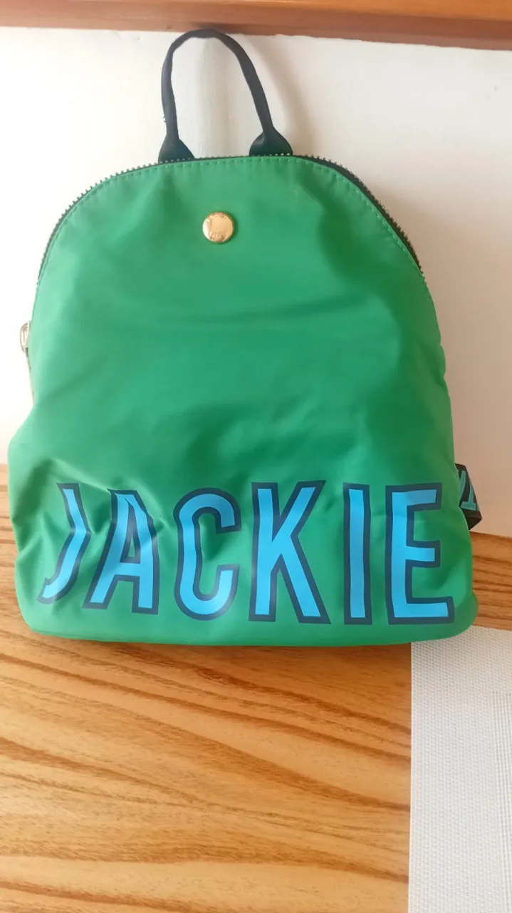 Mochila verde vibrante con el nombre "JACKIE" en letras grandes y celestes en el frente. Las tiras de la mochila también tienen el nombre "JACKIE SMITH" y estrellas estampadas. Ideal para el día a día o para llevar tus esenciales con estilo.