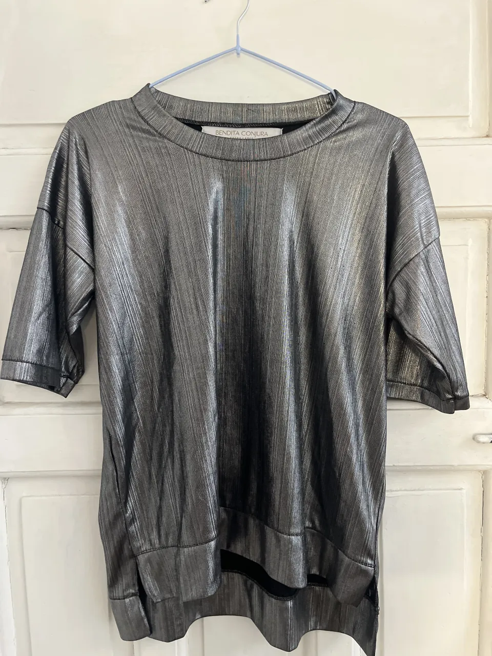 Remera plateada con textura de líneas verticales finas. Cuello redondo y mangas cortas a la altura del codo. Dobladillo inferior curvo.