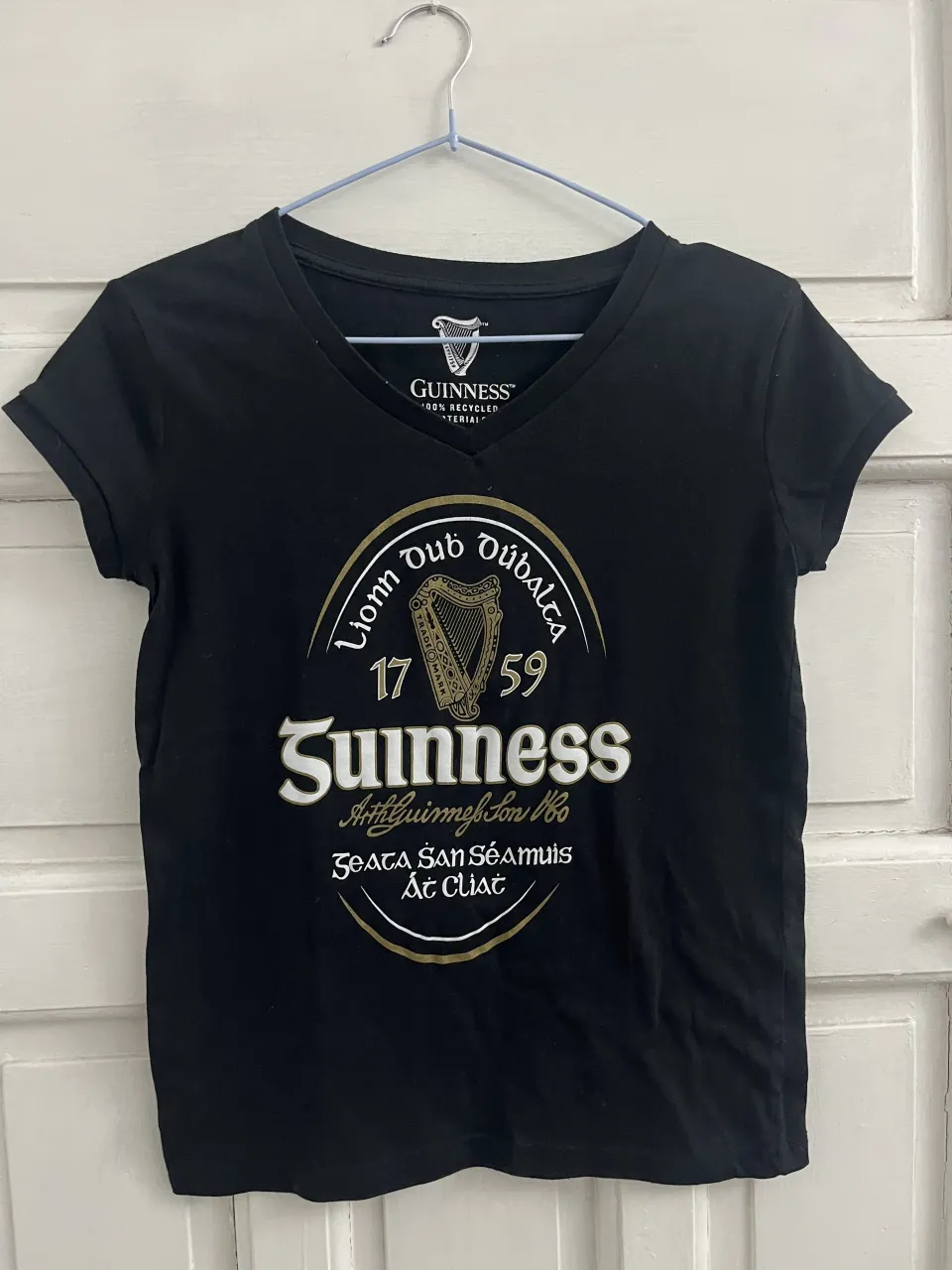 Remera negra de manga corta con cuello en V. Estampado frontal con el logo de Guinness y detalles dorados y blancos.