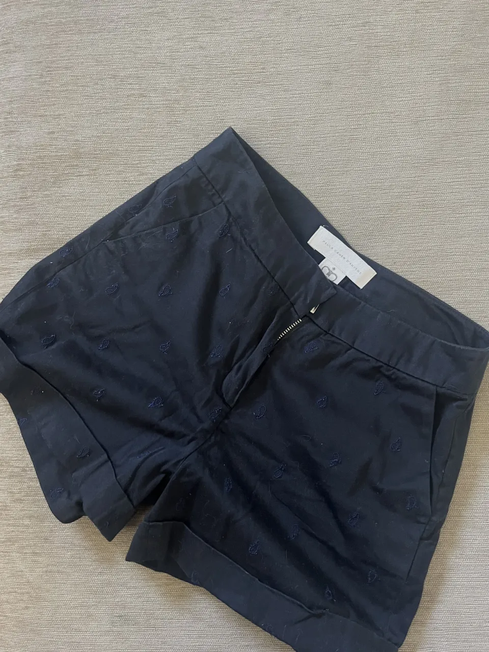 Shorts de gabardina azul marino con pequeños bordados de hojas en tono más oscuro. Tienen bolsillos delanteros y traseros, y el dobladillo de las piernas está doblado.