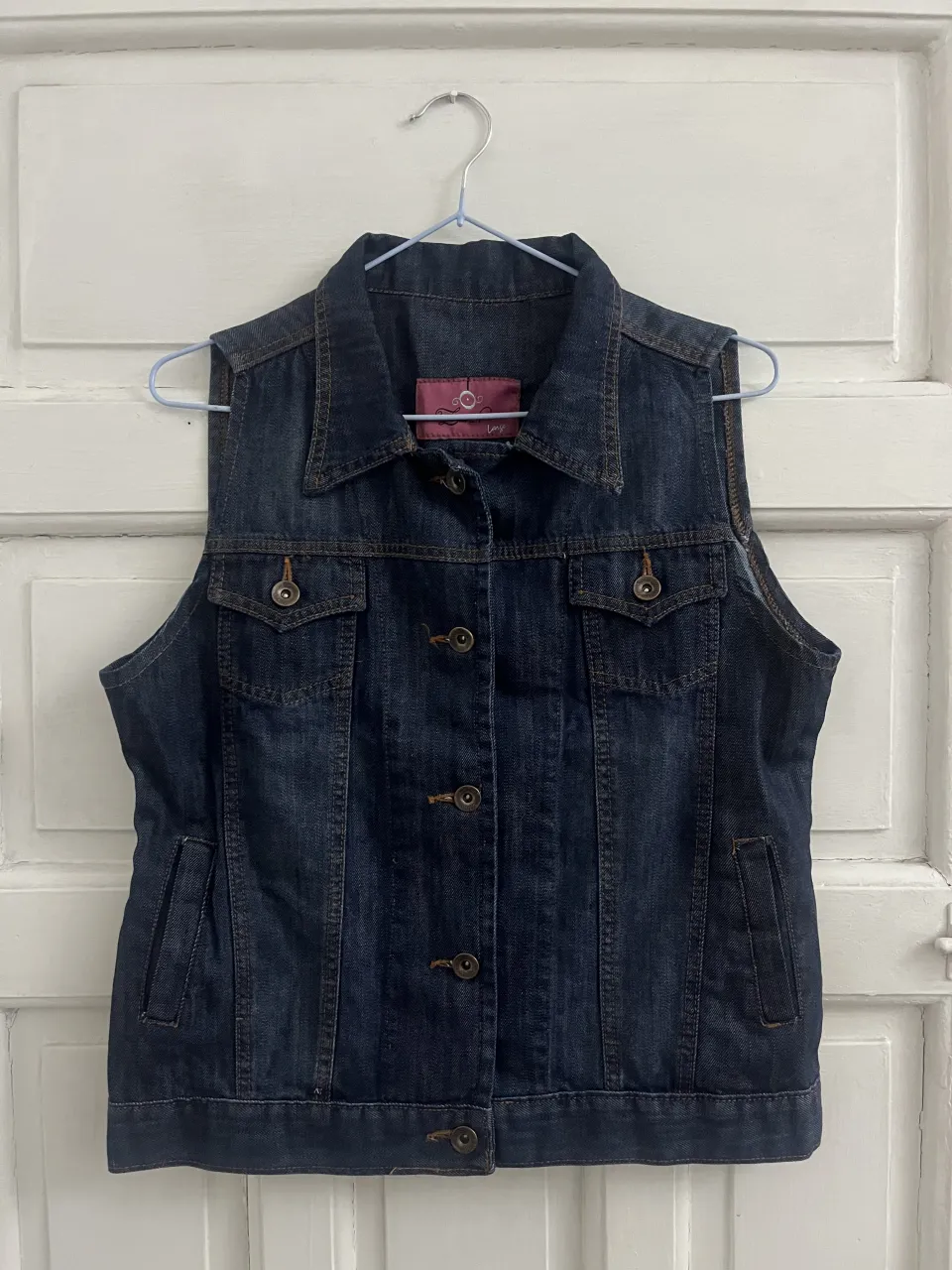 Chaleco de jean azul oscuro, con cuello de camisa, botones al frente y bolsillos con solapa en el pecho. Posee bolsillos laterales.