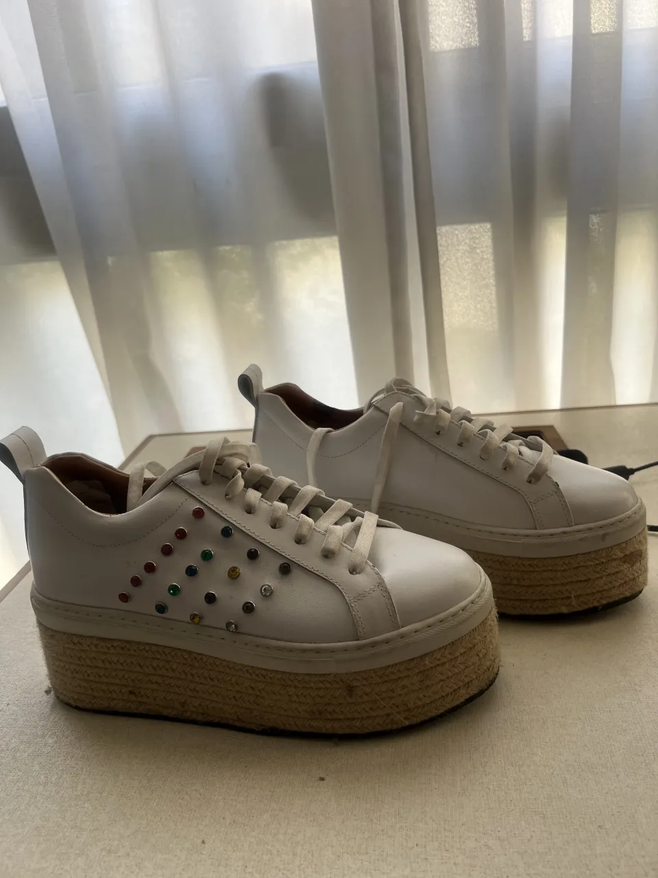 Zapatillas blancas de plataforma con detalle de tachas de colores en los cordones. La suela es de yute.