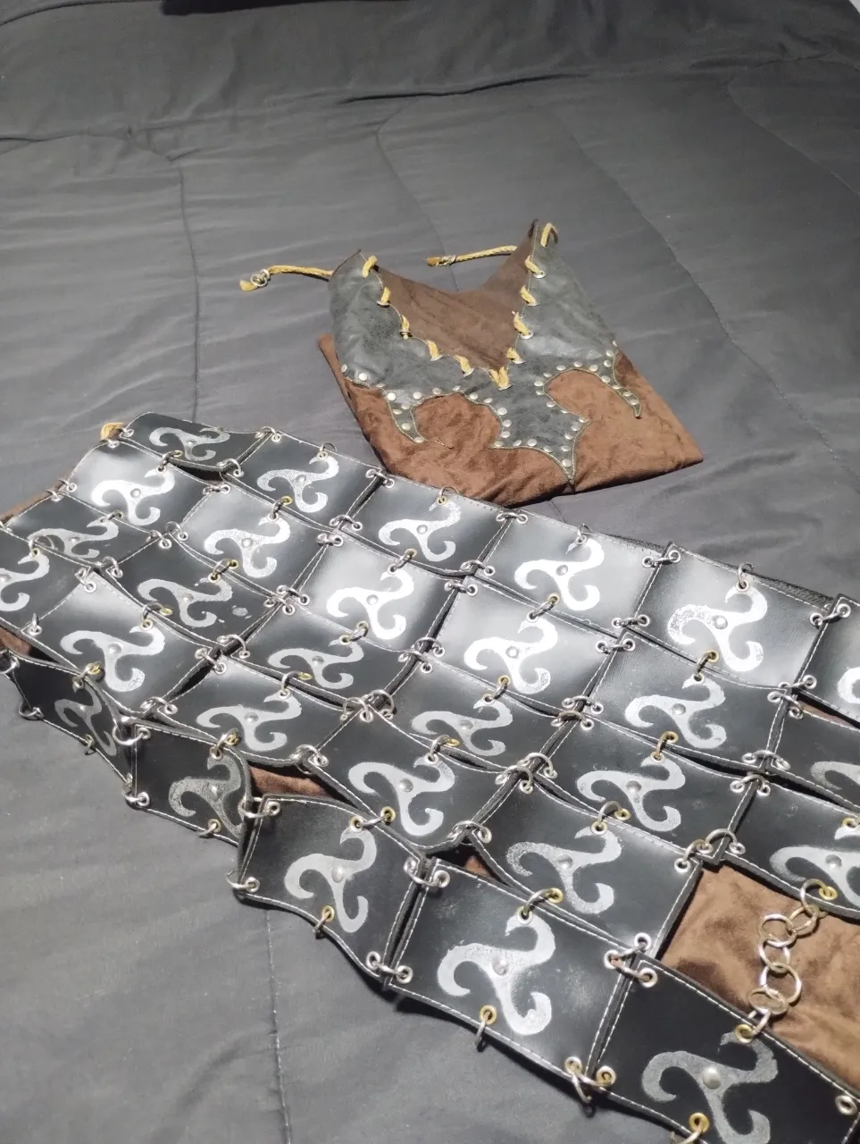 Conjunto de vestimenta estilo medieval o fantasía, TOTALMENTE ARTESANAL,ideal para eventos temáticos, cosplay o festivales. Incluye un corpiño o túnica sin mangas de color marrón con detalles de cuero negro y tachas plateadas, y una falda o delantal con placas cuadradas negras adornadas con un diseño plateado en espiral. El conjunto se cierra con cordones rústicos y tiene detalles de cadenas metálicas.