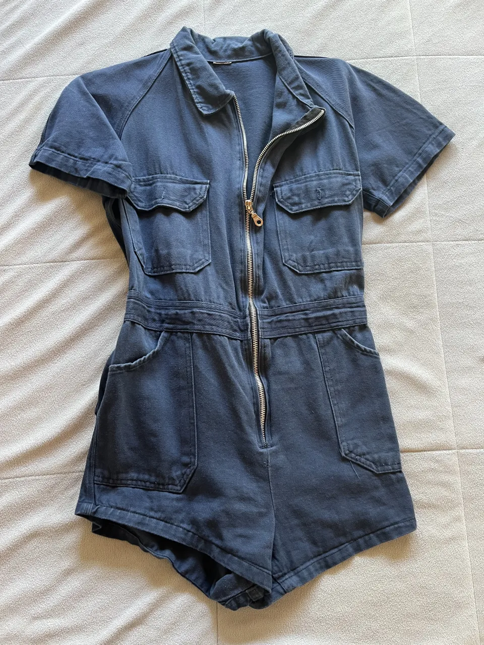 Mono corto de gabardina azul marino con cuello camisero, mangas cortas y cierre metálico al frente. Posee bolsillos superiores con solapa y bolsillos inferiores tipo ojal. Cintura con elástico fruncido en la espalda y detalle de cinturón fino. Bolsillos traseros con solapa.