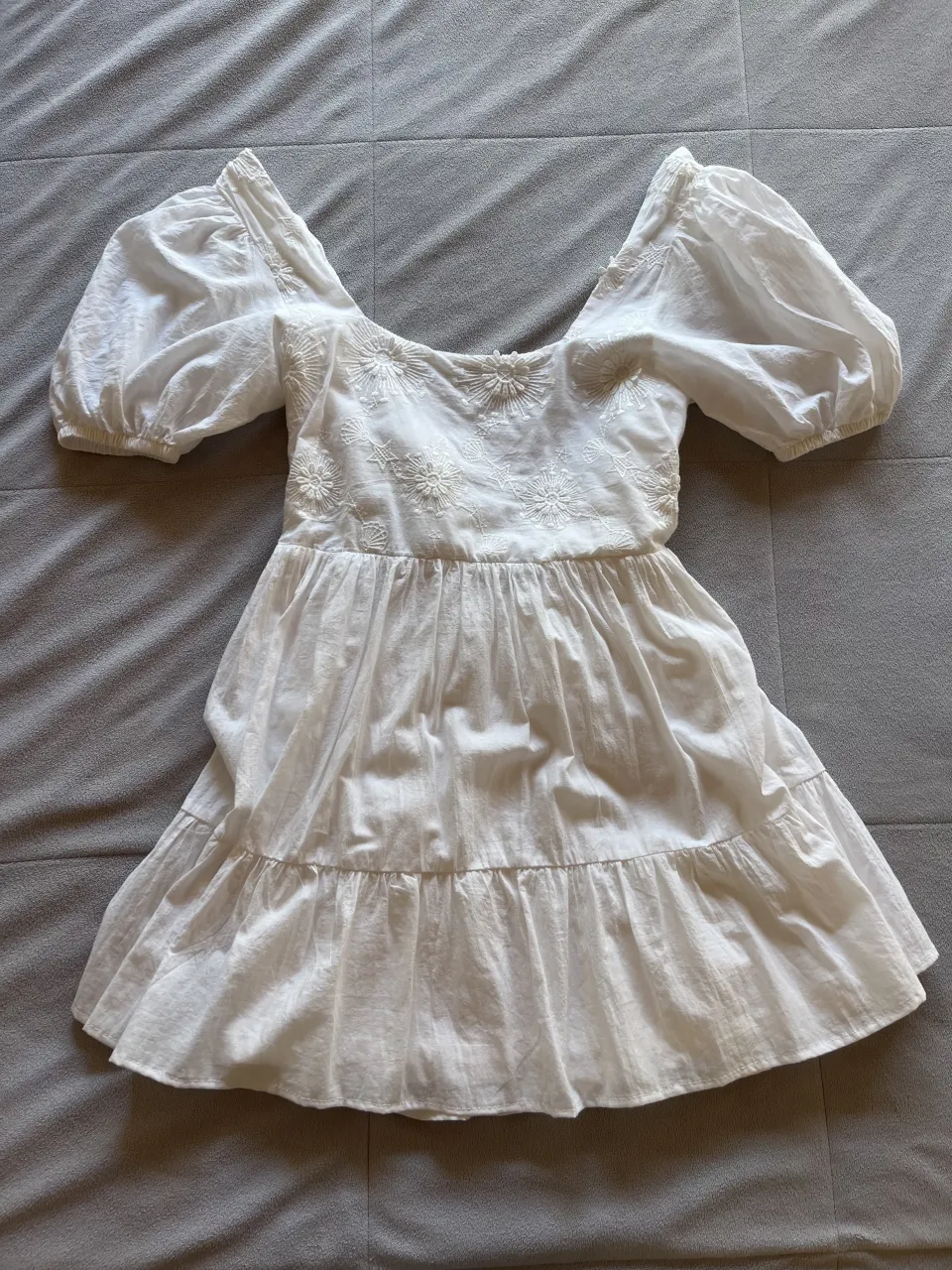 Vestido blanco corto con mangas abullonadas y escote cuadrado. Posee bordados florales en la parte superior y un lazo en la espalda con un recorte. La falda es con volados.