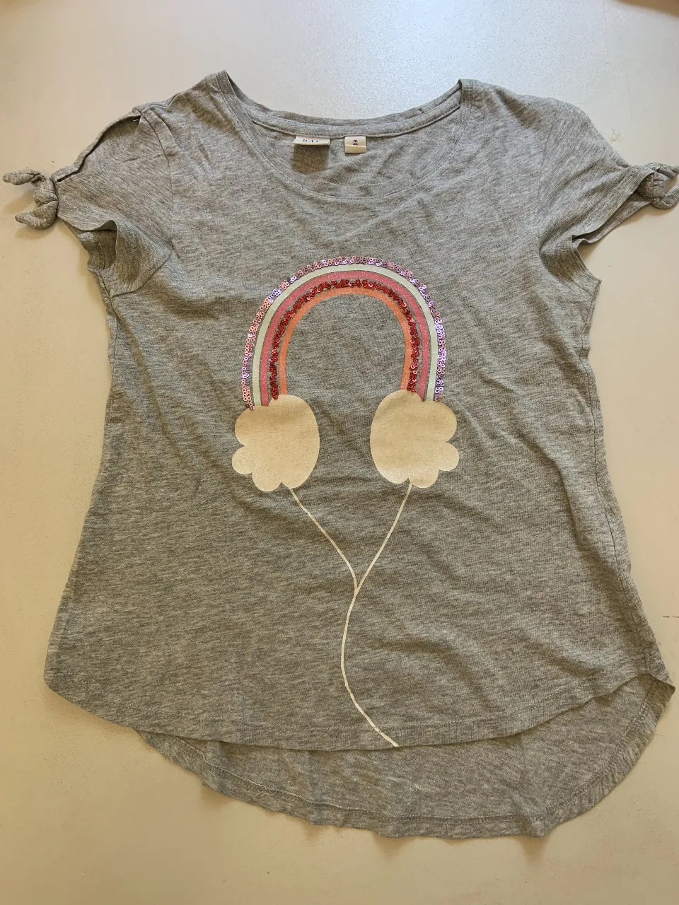 Remera gris de manga corta para niña, con diseño de arcoíris y auriculares estampados con lentejuelas. Mangas con detalle de lazo. Ideal para un look casual y divertido.