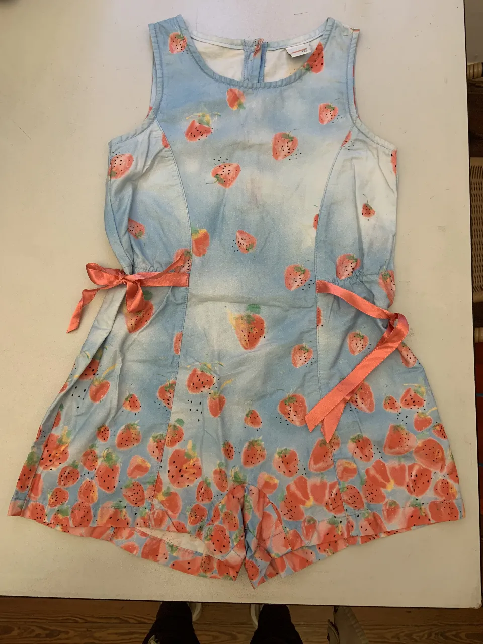 Mono corto de niña con estampado de frutillas sobre un fondo celeste. Cuenta con lazos color coral en la cintura y botones en la espalda. Ideal para el verano.