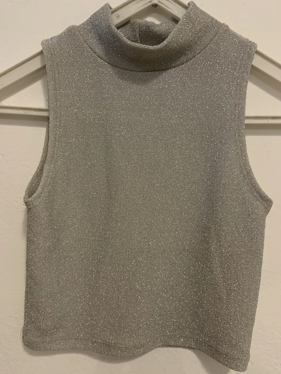 Top sin mangas de mujer, de cuello alto y corte crop. Confeccionado en tela brillante plateada, ideal para un look de fiesta o salida.