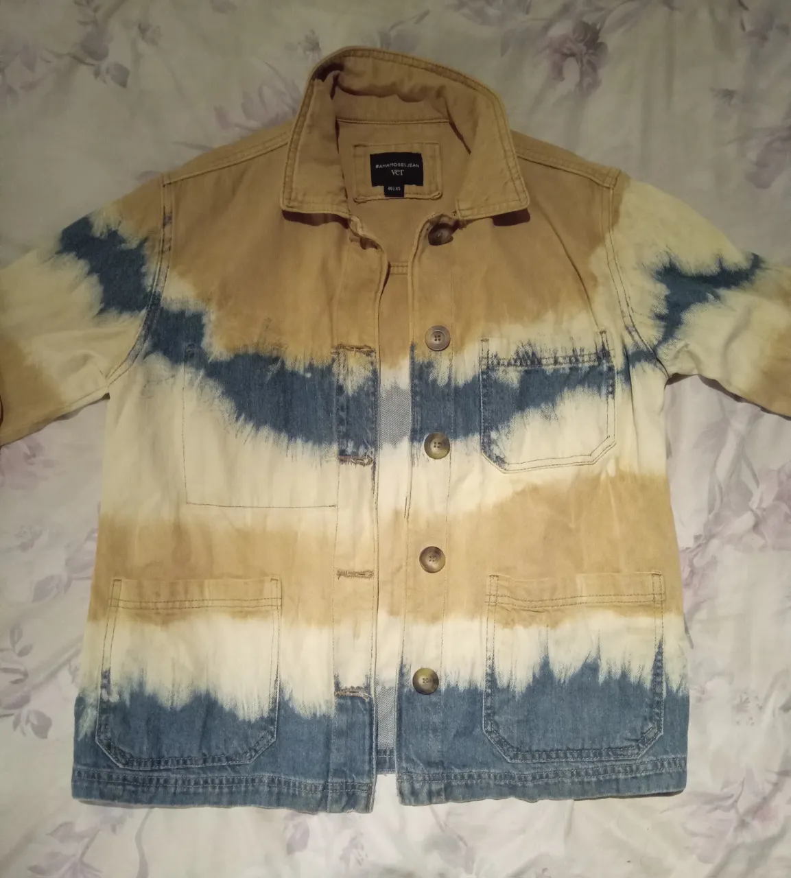 Campera de jean con diseño tie-dye en tonos beige, blanco y azul. Posee cuello camisero, botones al frente y bolsillos con solapa en el pecho y en la parte inferior.