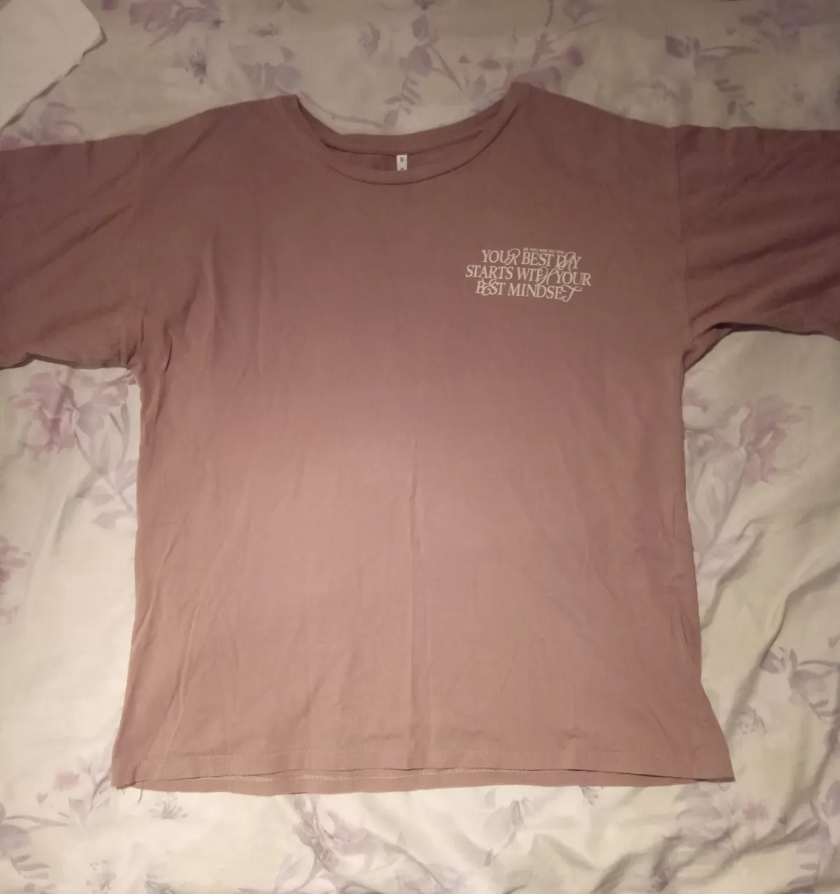 Remera de color rosa con estampado en la parte frontal que dice "YOUR BEST DAY STARTS WITH YOUR BEST MINDSET". Tiene un diseño de cuello redondo y mangas cortas. La prenda es sencilla y sin detalles adicionales.