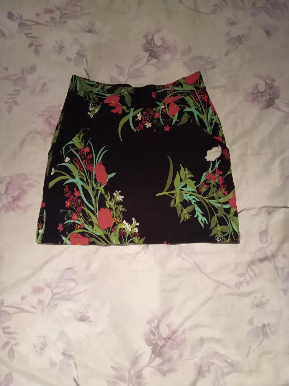 Pollera corta negra con estampado floral vibrante. Presenta flores rojas y verdes con detalles en blanco y verde menta, creando un diseño llamativo y fresco. Ideal para un look casual y femenino.