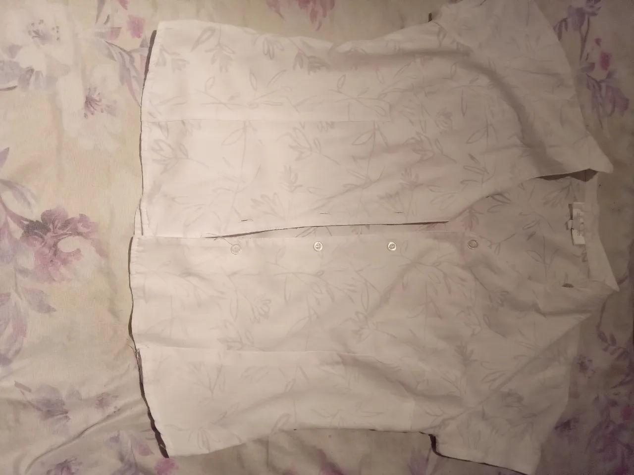 Camisa blanca de manga corta con un delicado estampado floral en tonos grises. Posee botones al frente y un corte clásico, ideal para un look casual y fresco.