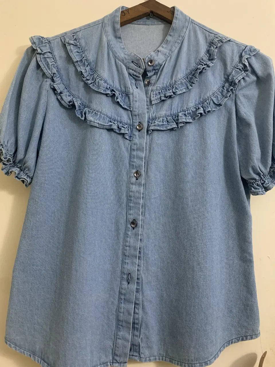 Blusa de jean azul claro con cuello alto y volados en el cuello y mangas cortas. Cierre con botones al frente.