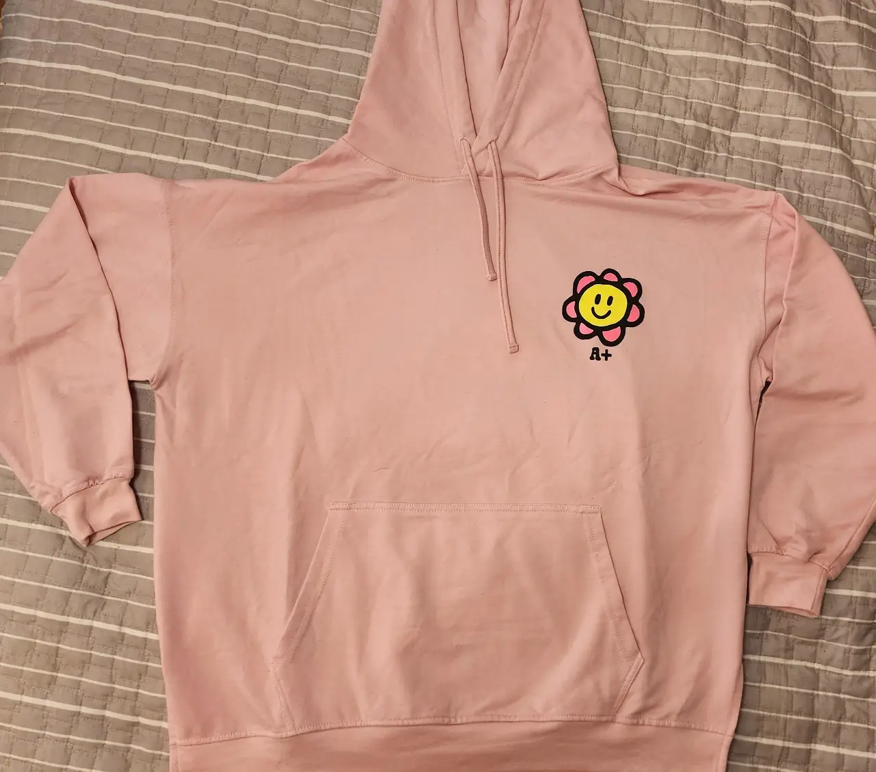 Buzo canguro rosa con estampado de flor sonriente y logo A+. Ideal para un look casual y cómodo.