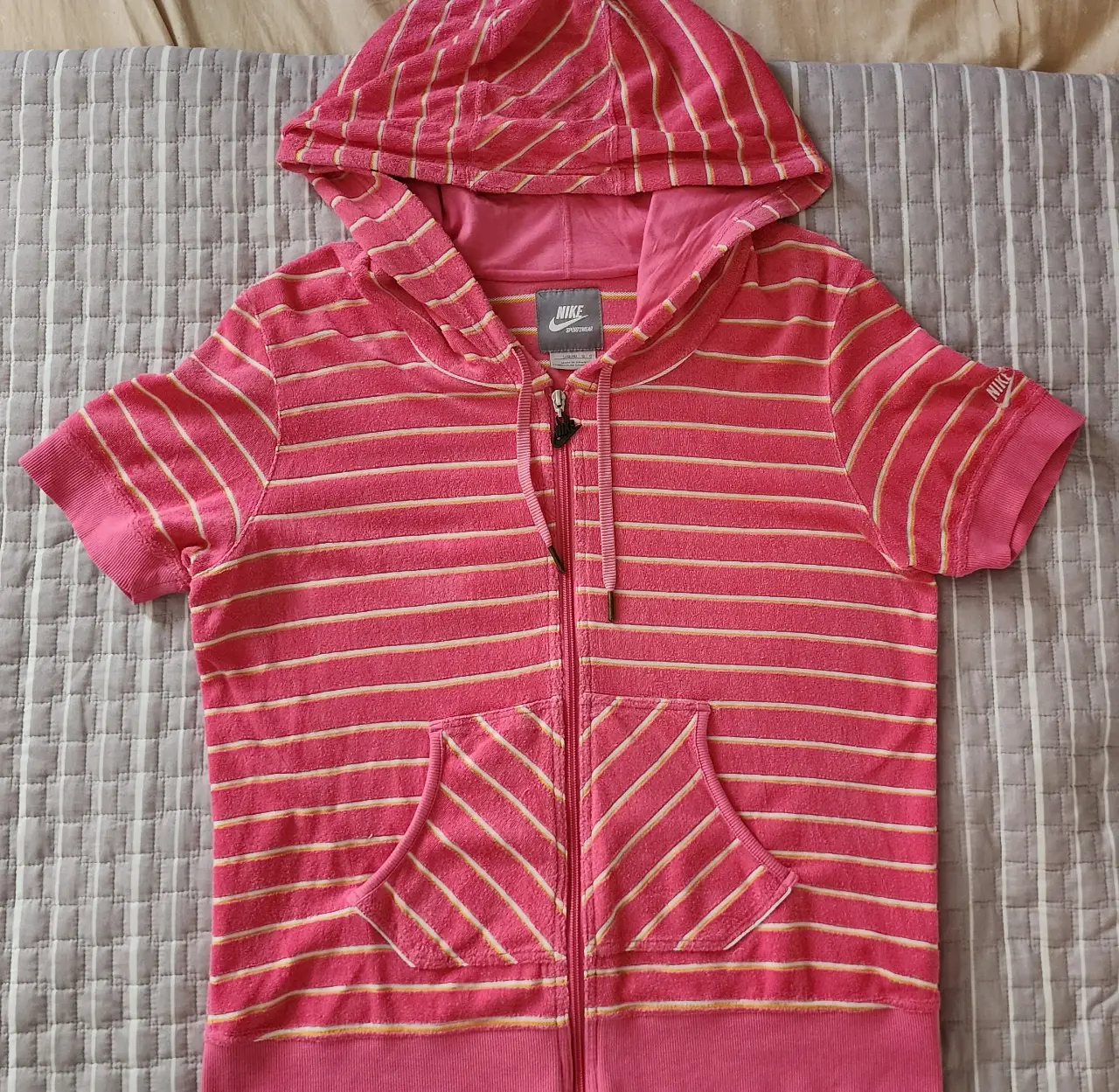 Buzo con cierre y capucha, de manga corta, con rayas horizontales rosas y blancas. Con bolsillo frontal y logo de Nike. Ideal para un look casual y deportivo.