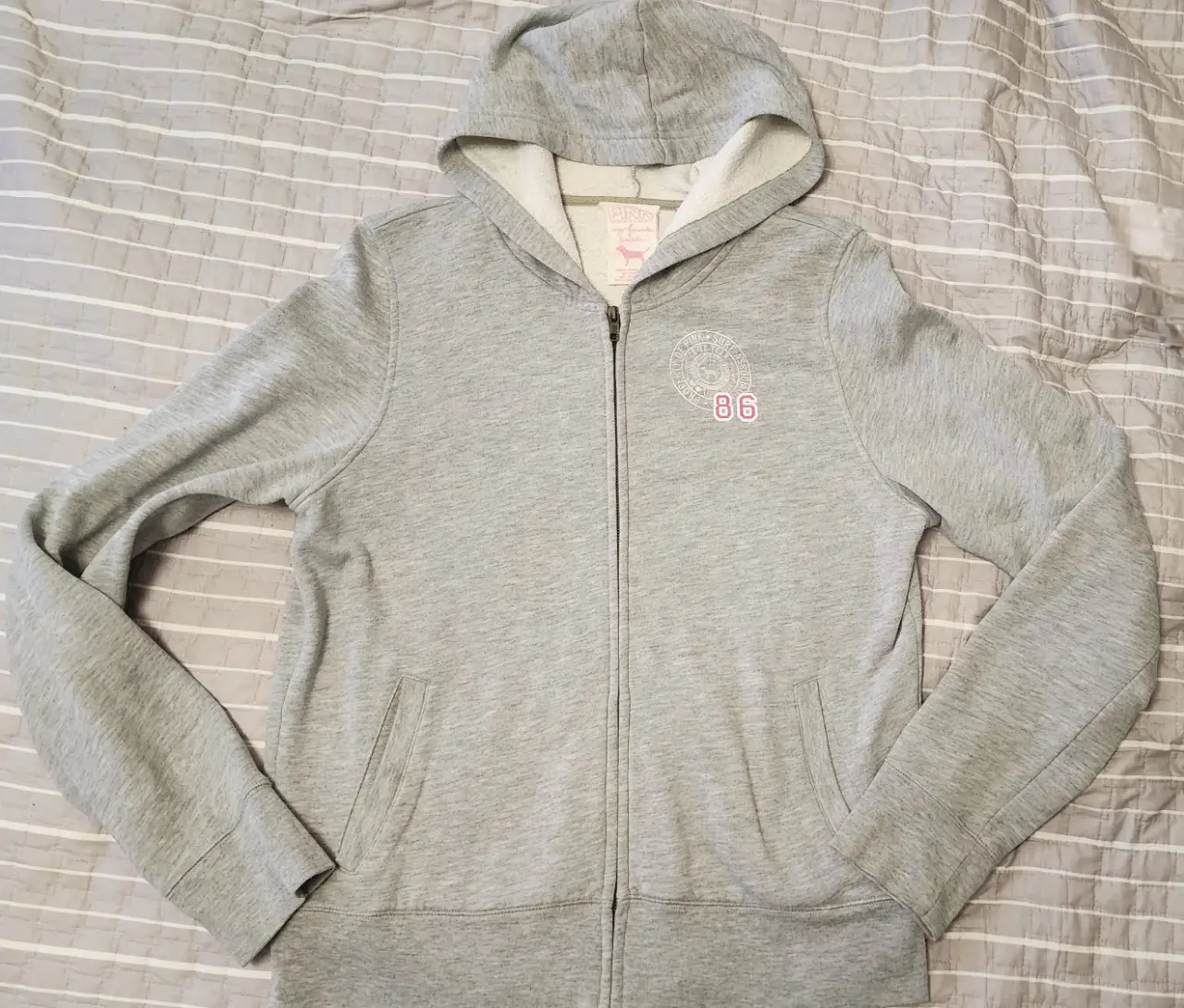 Buzo con cierre y capucha de la marca Pink, color gris con detalles en rosa. Tiene un estampado en el frente y la inscripción "Team Pink" en la espalda. Talle L.