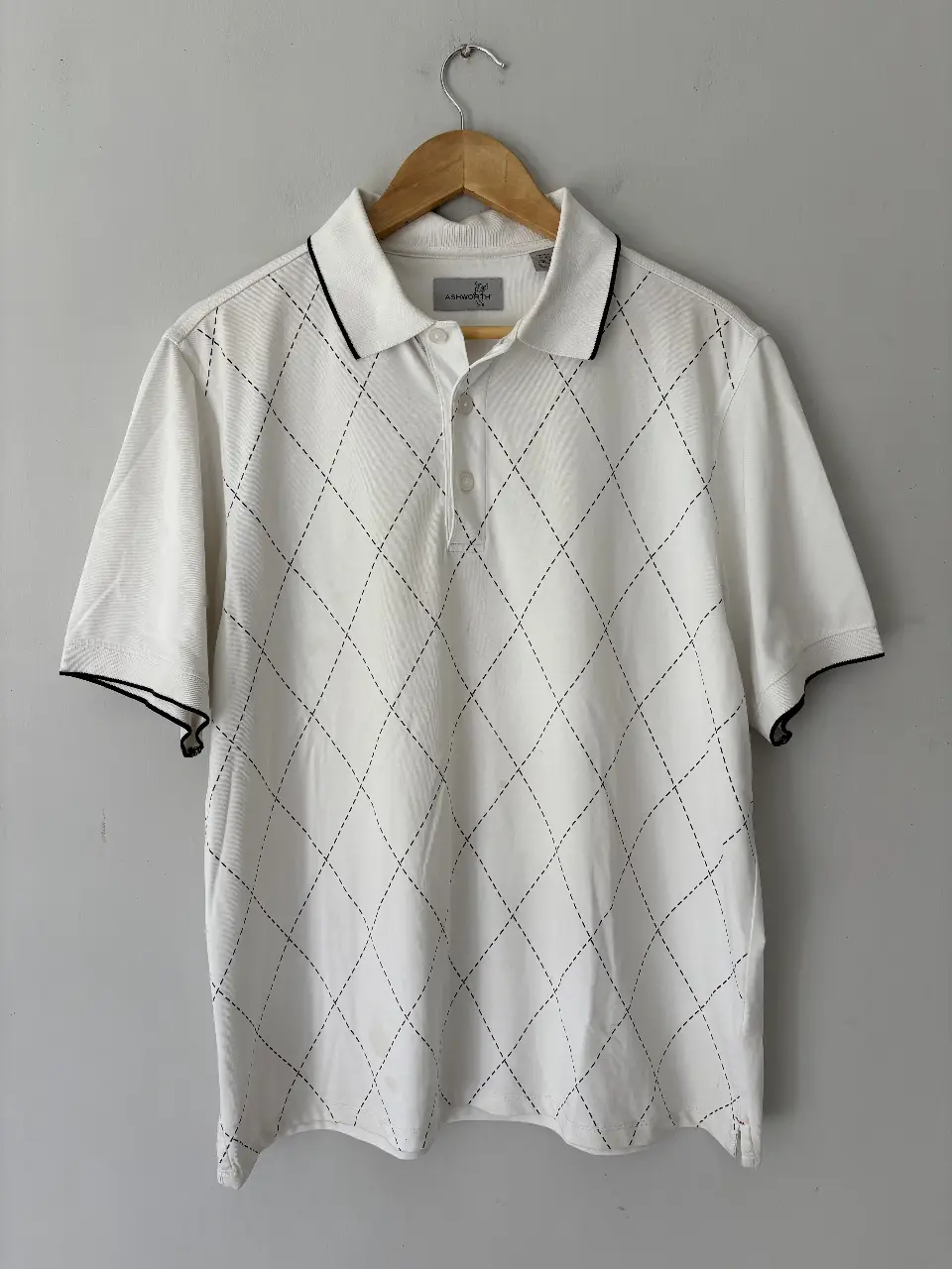 Remera polo Ashworth blanca con estampado de rombos en negro. Cuello y mangas con detalles en negro. Talle M.