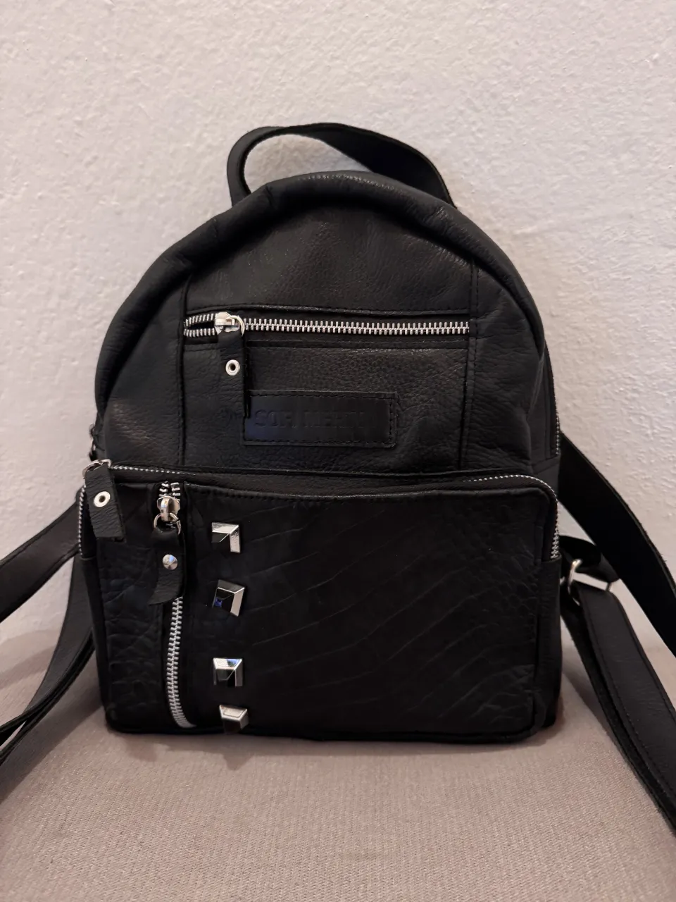 Mochila negra de cuero, con tachas plateadas y detalles de textura. Tiene varios bolsillos con cierre y un interior forrado en color violeta. Ideal para un look rockero o casual. medidas: 30x26x6cm