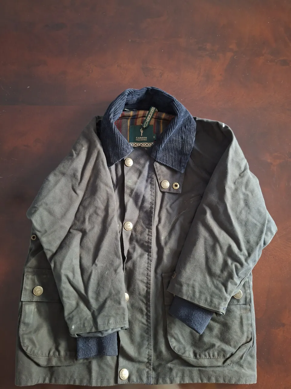 Campera de tela impermeable, color verde oscuro, con cuello de pana azul marino. Posee botones a la vista y bolsillos frontales con solapa. Puños de tejido acanalado.