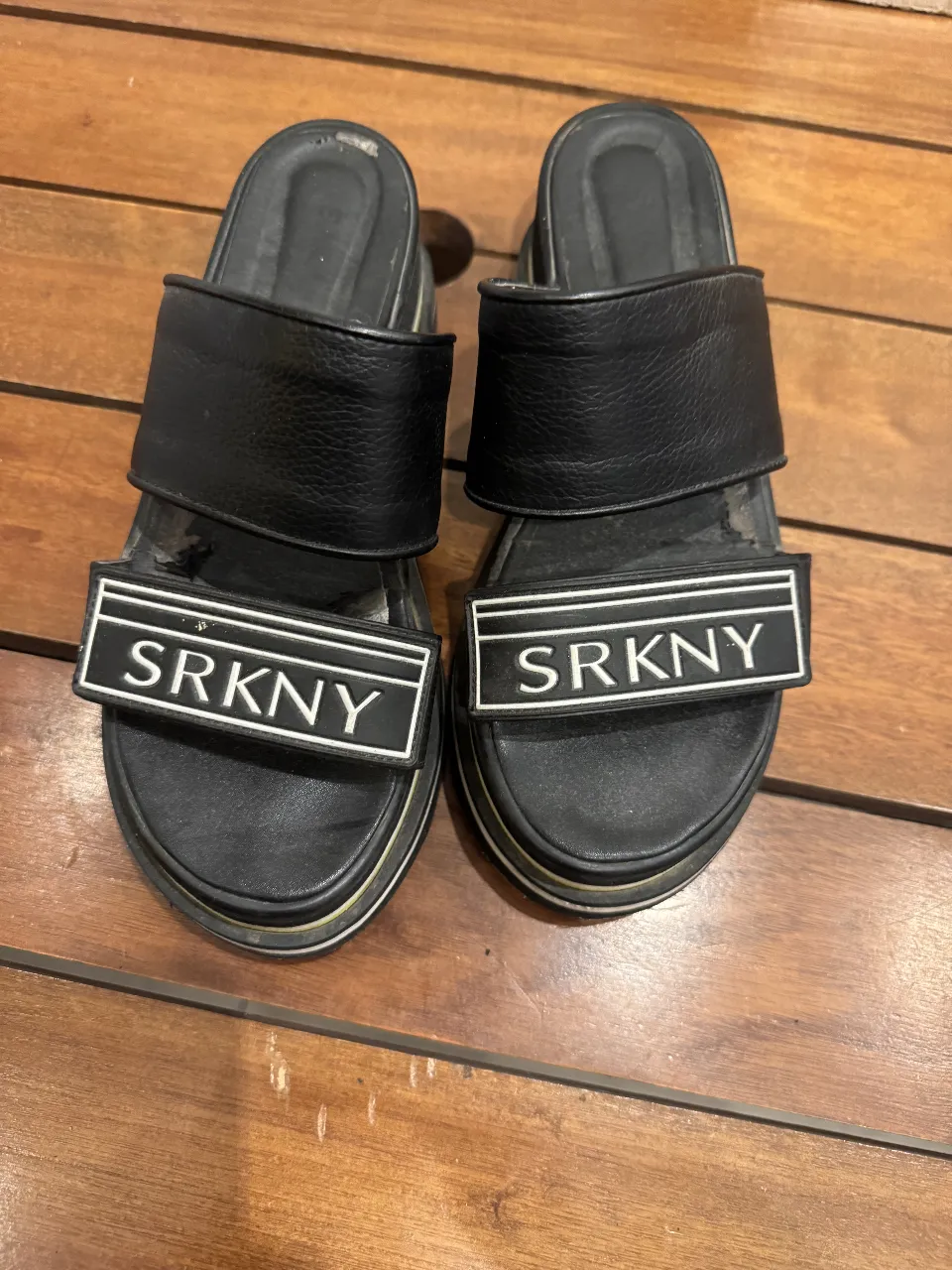 Sandalias negras con plataforma y detalles en blanco. Tienen una tira ancha en la parte superior con la inscripción "SRKNY" en blanco. Ideales para un look casual y moderno.