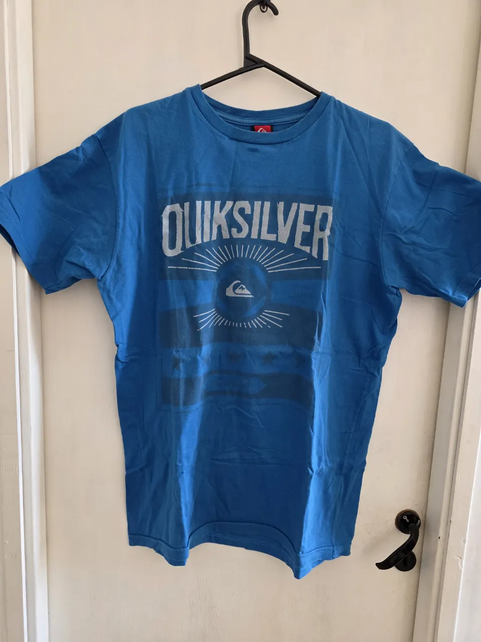 Remera azul de manga corta con estampa de la marca Quiksilver en blanco y gris. La estampa incluye el logo de la marca y el nombre.