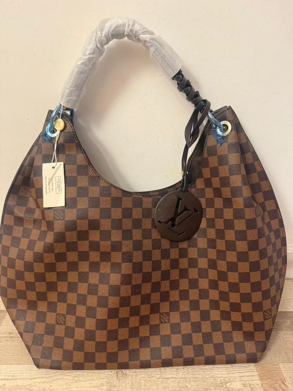 Cartera Louis Vuitton modelo hobo, confeccionada en lona Damier Ebene. Diseño con asa superior enrollada, dije con logo LV y cierre superior. Interior espacioso con bolsillo con cierre. Símil 