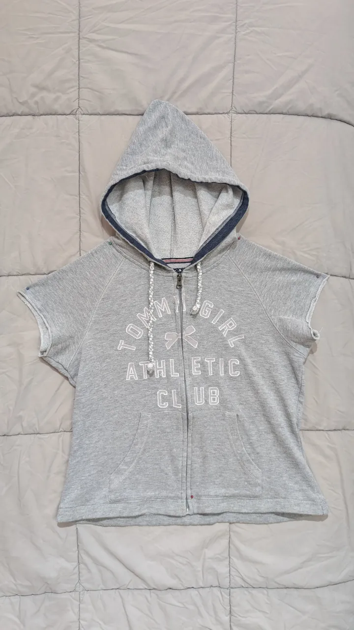 Buzo con capucha y cierre completo, de mangas cortas, en color gris melange. Posee estampa "Tommy Girl Athletic Club" en el frente y bolsillos tipo canguro. Ideal para un look casual y deportivo.