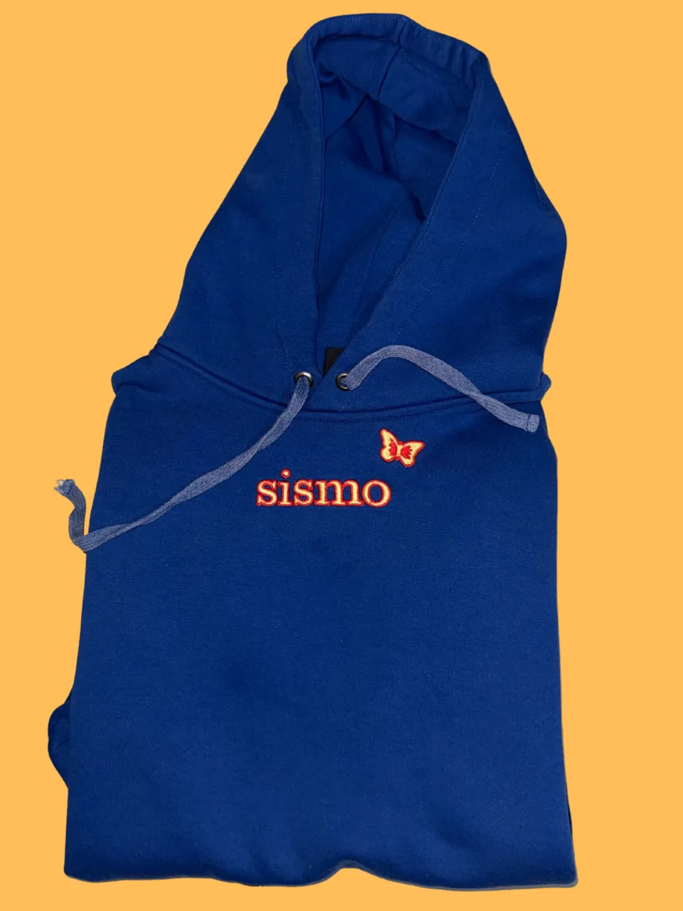 Buzo azul con capucha y cordones grises. Tiene un bordado de la palabra "sismo" en color rojo y una mariposa naranja con detalles rojos.