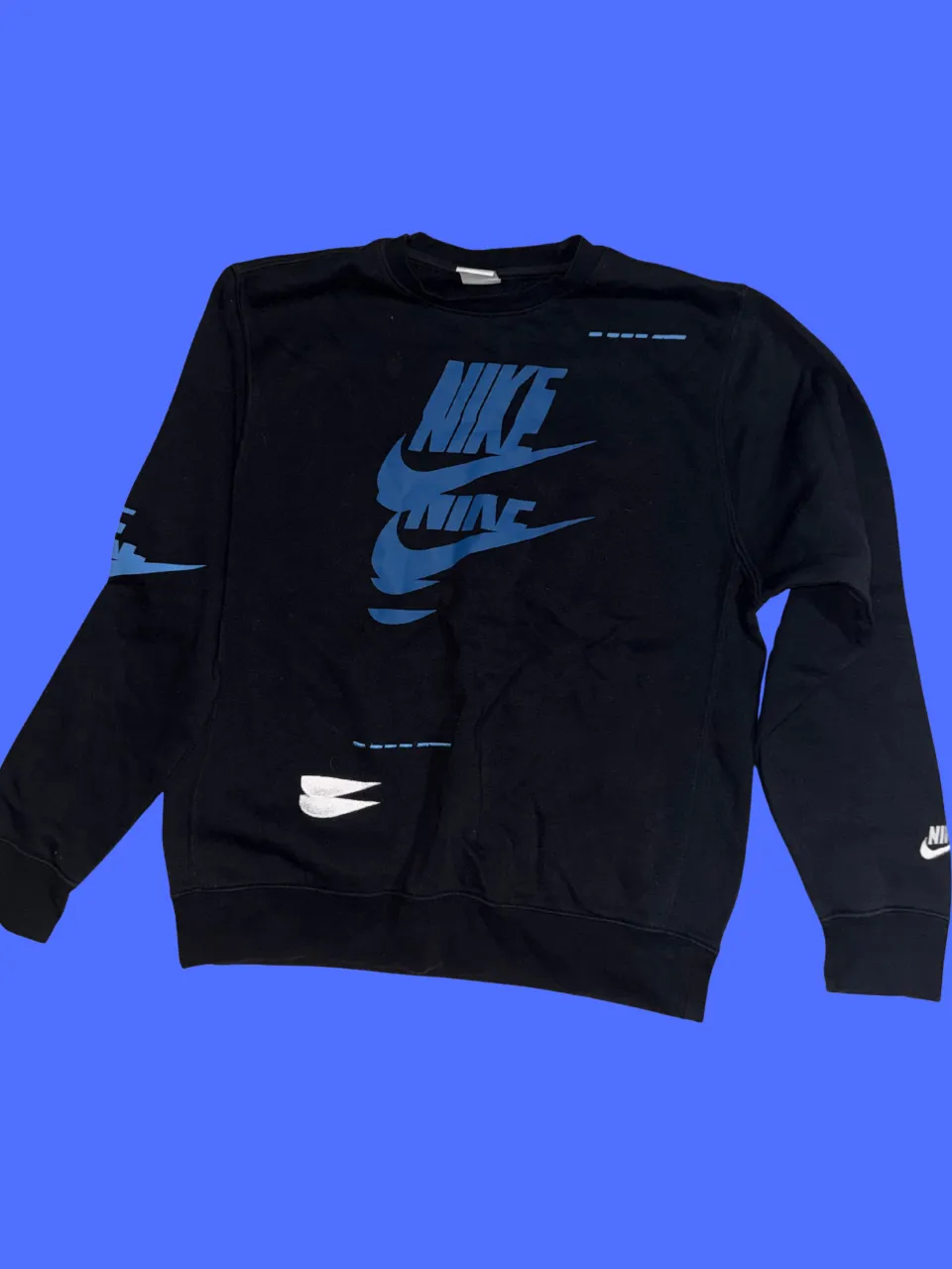 Buzo negro con estampa de Nike en frente y espalda. Estampa frontal en color azul con el logo de la marca y "NIKE" repetido. Estampa trasera con logo de Nike en blanco y azul. Mangas largas. Cuello redondo.