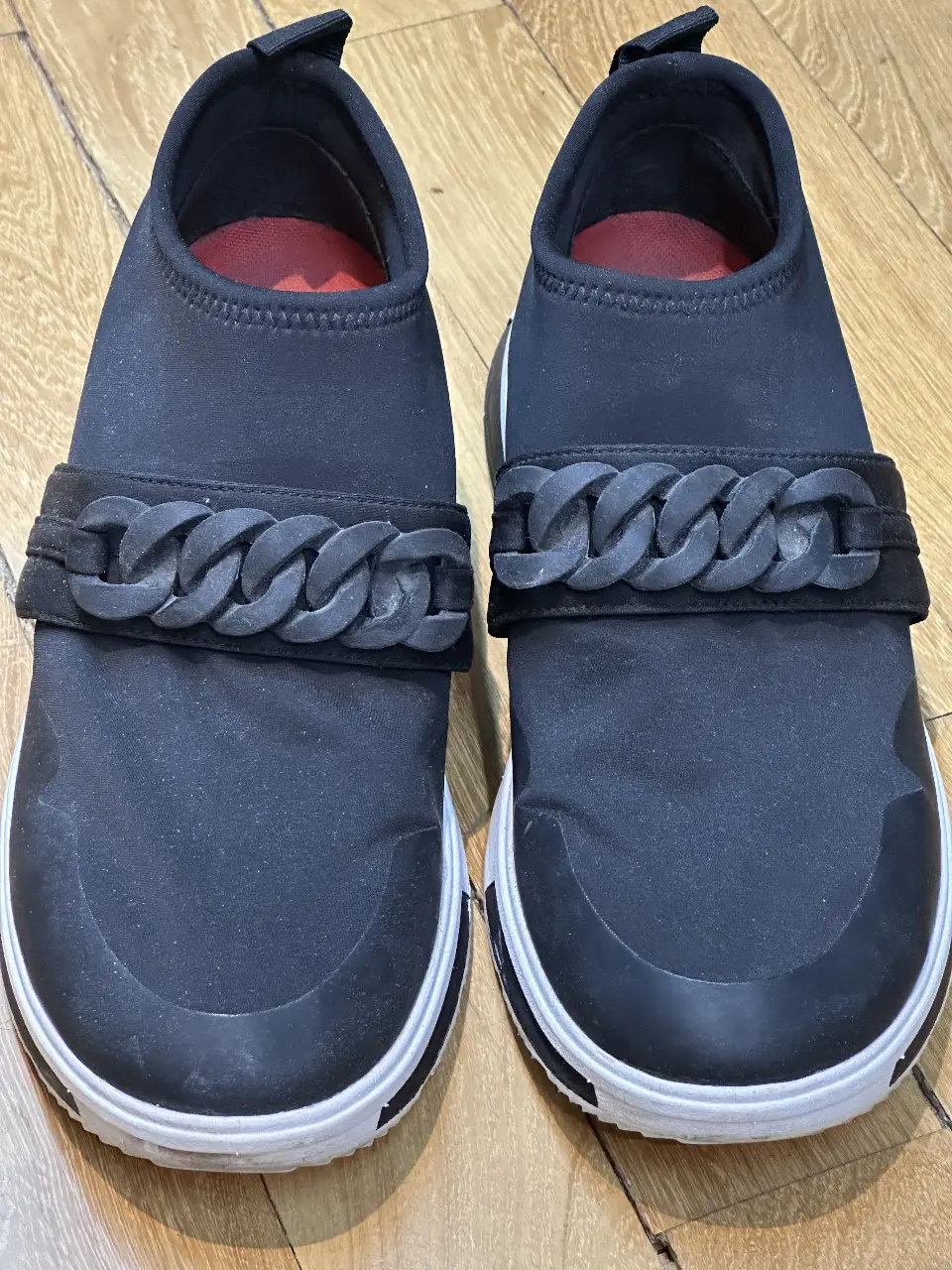 Zapatillas slip-on negras con detalle de cadena de goma en el empeine y suela blanca. Cómodas y modernas, ideales para un look casual.