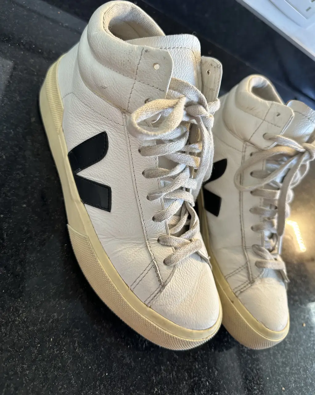 Zapatillas Veja V-10 de cuero blanco con el logo V en negro. Tienen un diseño de caña alta y suela de goma color crema. Ideales para un look casual y moderno.