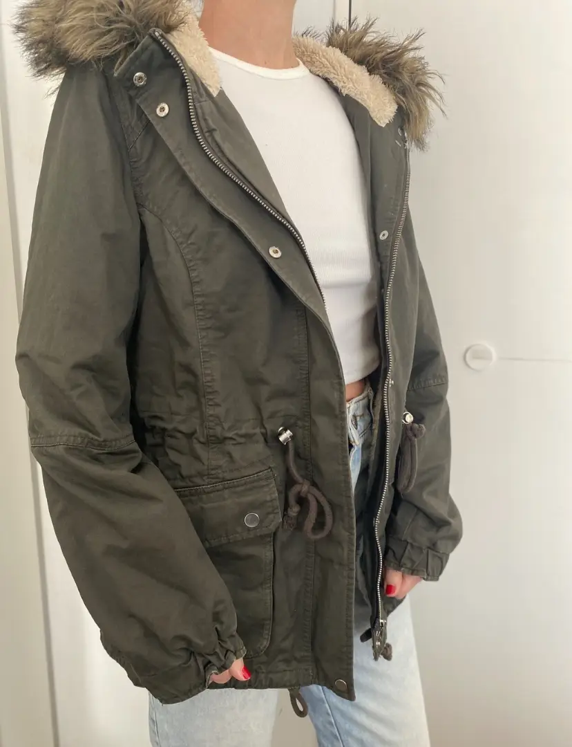 Campera tipo parka de mujer, color verde oscuro, con capucha con borde de piel sintética marrón. Interior forrado en corderito color crema. Cierre y botones frontales, bolsillos delanteros y ajuste en la cintura con cordón. Talle M. Marca H&M Divided.