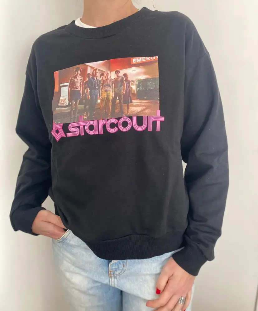 Buzo MARCA FALABELLA de cuello redondo negro con estampado de la serie Stranger Things y el logo de StarCourt en rosa. Ideal para un look casual y cómodo.