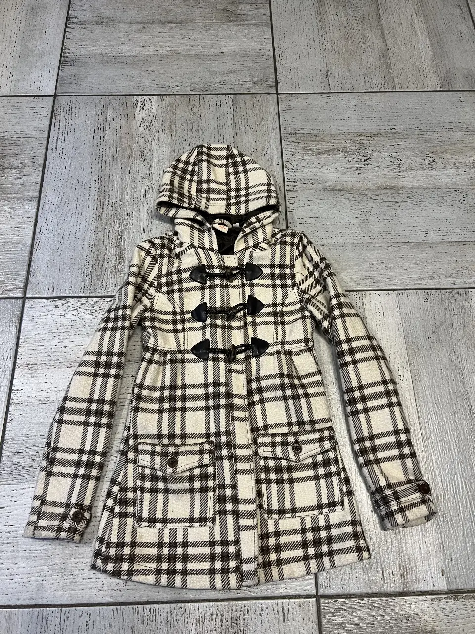Tapado con capucha y diseño a cuadros en tonos blanco y marrón. Cuenta con cierre de botones tipo duffle coat y bolsillos delanteros. Ideal para el invierno.