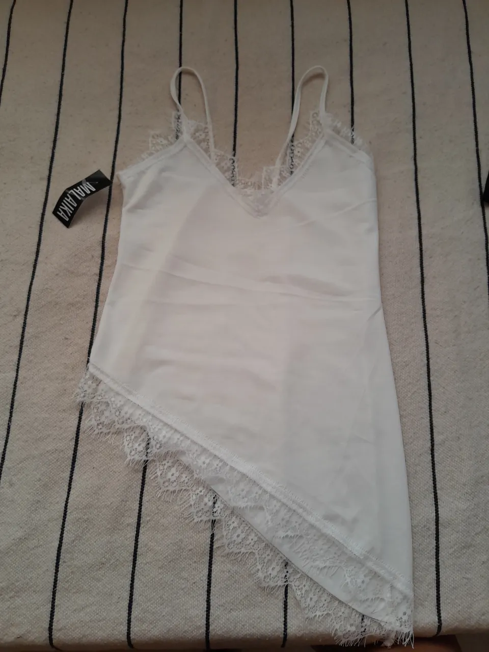 Top musculosa blanca con breteles finos y detalles de encaje en el escote y en el borde inferior asimétrico.