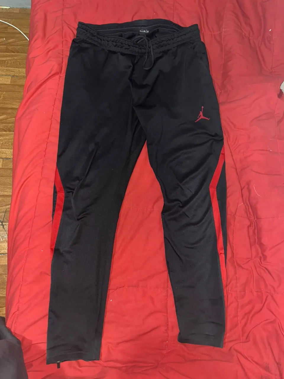 Pantalón deportivo Jordan negro con detalles rojos en los laterales y logo de Jordan bordado en rojo en la parte delantera. Posee cordón de ajuste en la cintura y cierre en los tobillos.