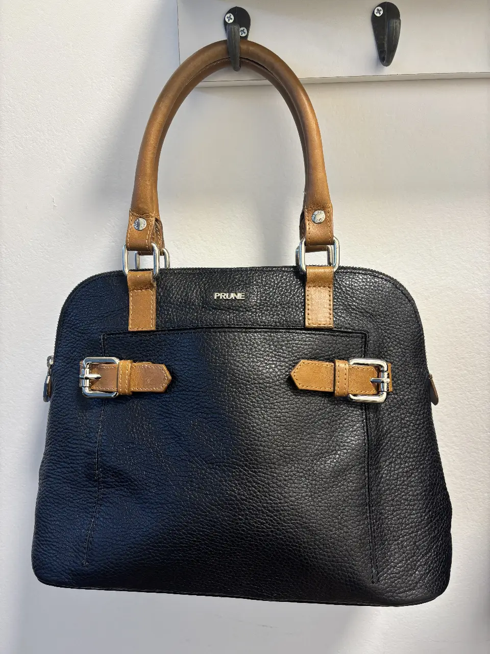 Cartera PRUNE de cuero negro con detalles marrones. Diseño elegante con manijas superiores y correa para colgar. Ideal para llevar tus esenciales con estilo.