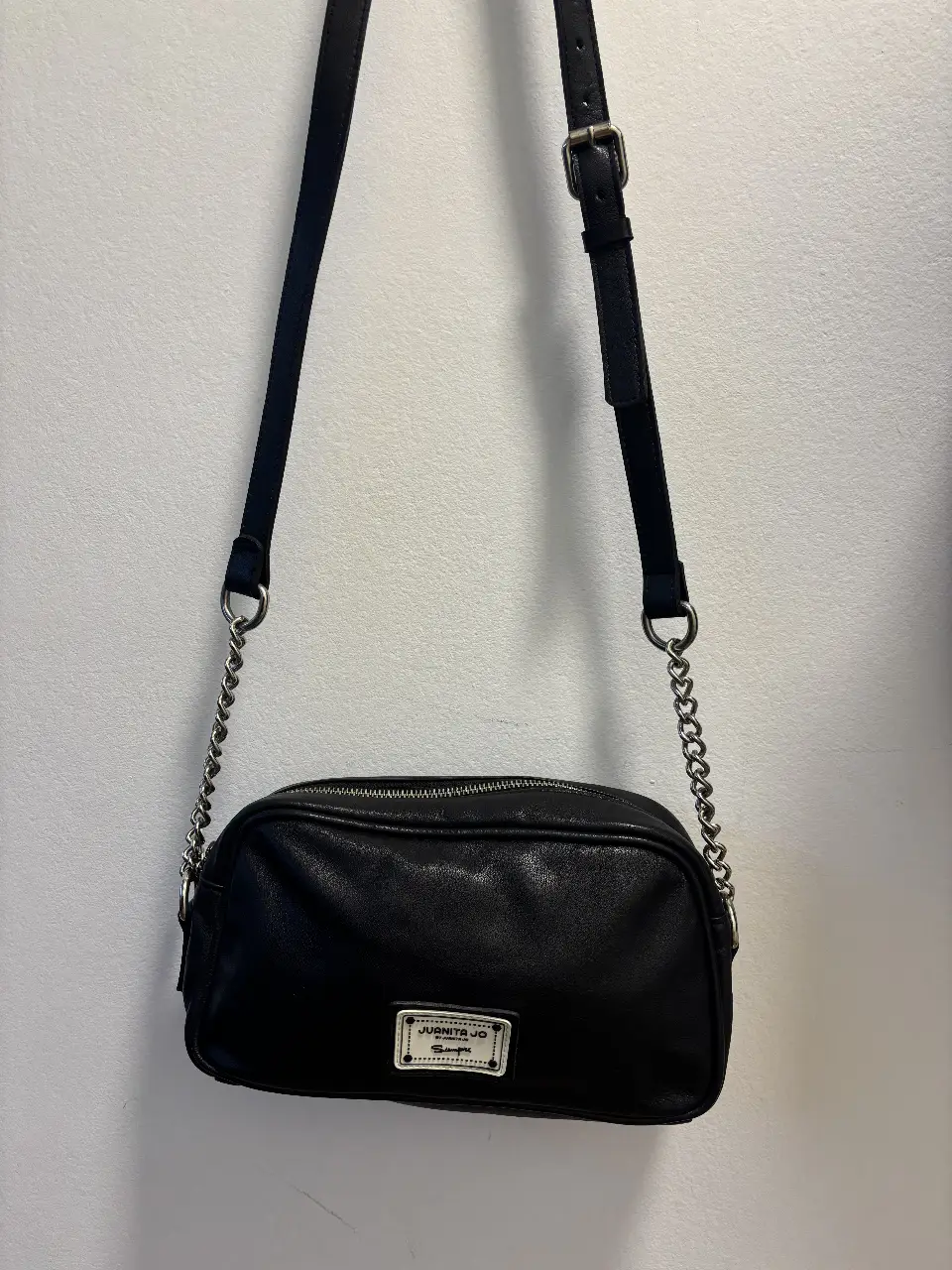 Bandolera negra de cuero sintético con correa ajustable y detalles de cadena. Ideal para un look casual y moderno.