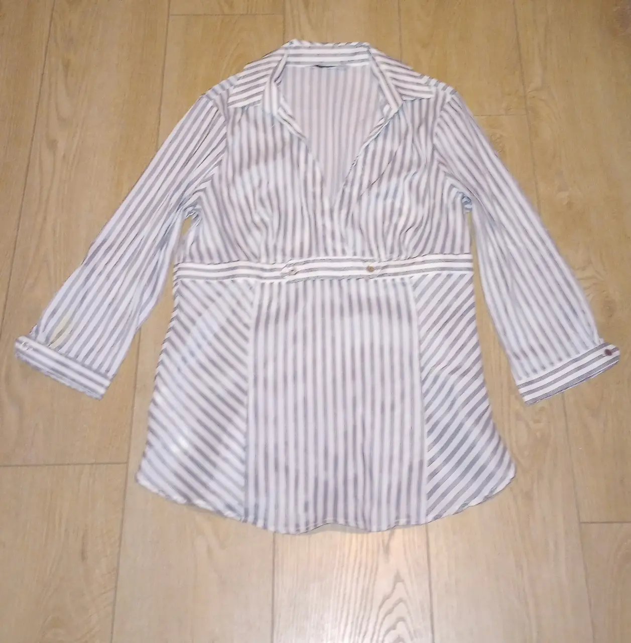 Blusa a rayas verticales en blanco y gris, con mangas largas y detalle de ajuste en la cintura. Ideal para un look elegante y versátil.