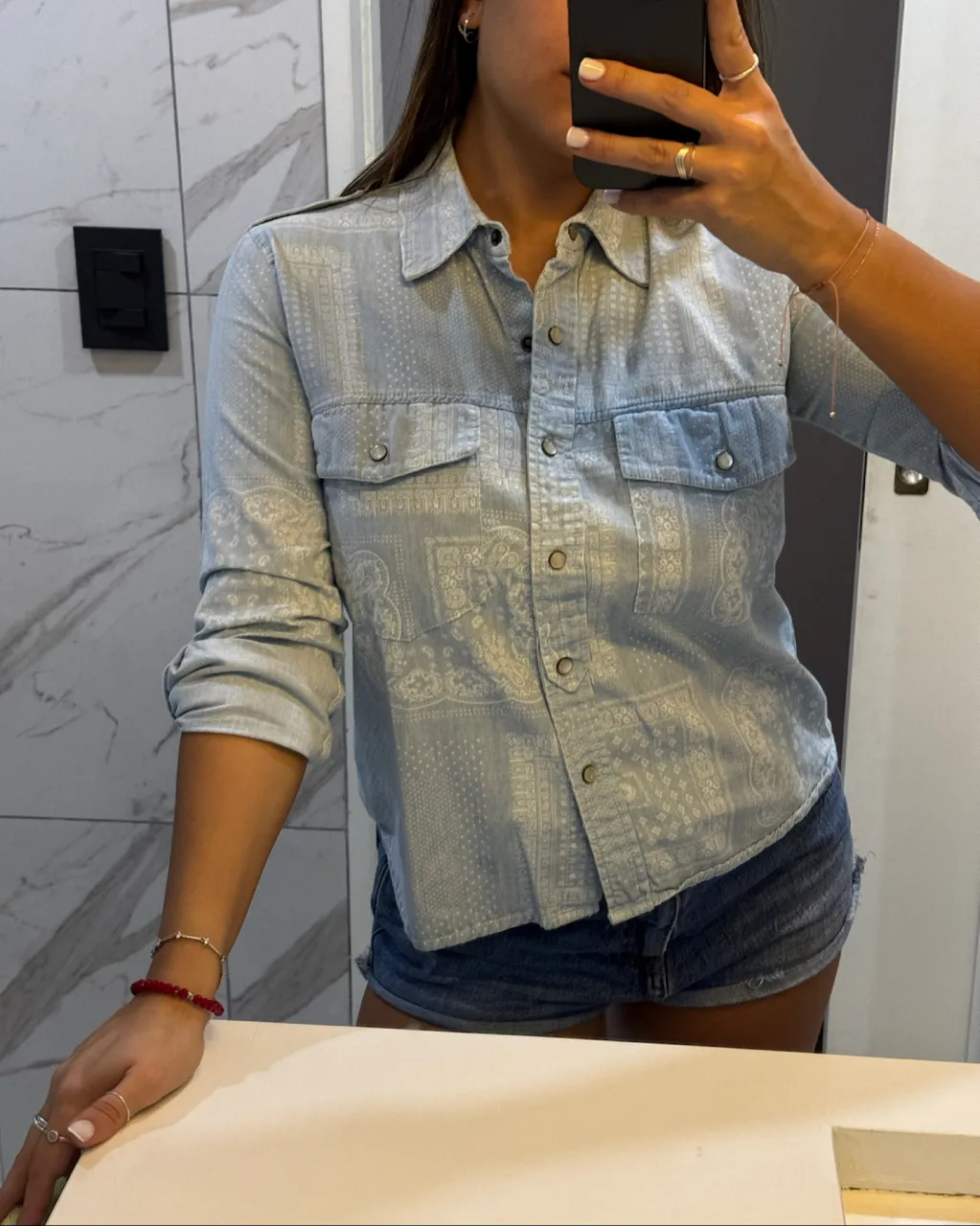 Camisa de jean celeste con estampado blanco de motivos geométricos y florales. Posee cuello, mangas largas con puños y botones, dos bolsillos en el frente con solapa y botones, y botones en el frente para cerrar.