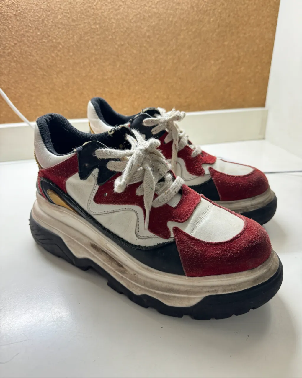 Zapatillas deportivas con cordones, suela gruesa y diseño de paneles en blanco, rojo, negro y detalles en dorado.