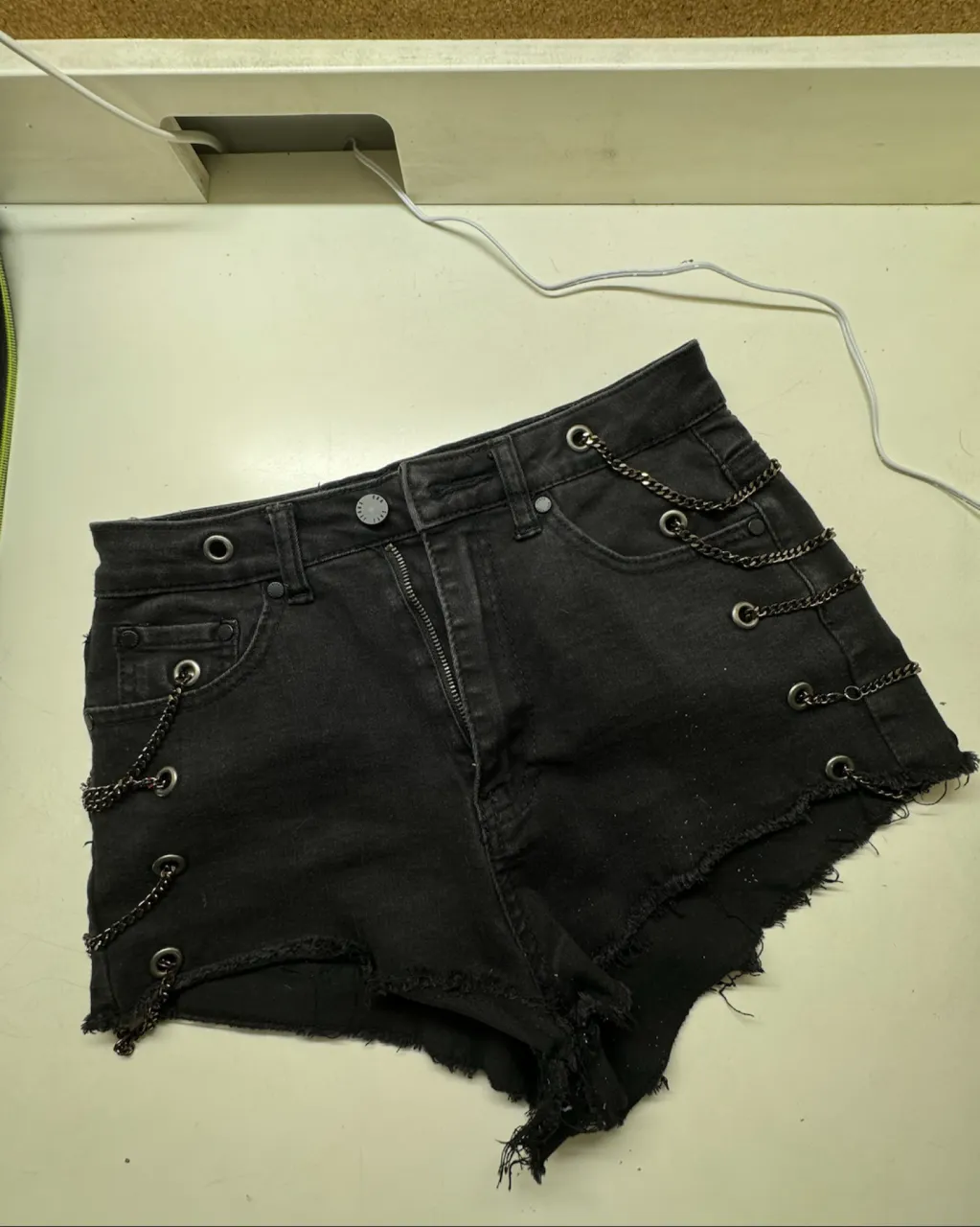 Shorts de jean negros con roturas y cadenas metálicas decorativas en los laterales. Tienen detalles de ojales metálicos y el borde inferior deshilachado.