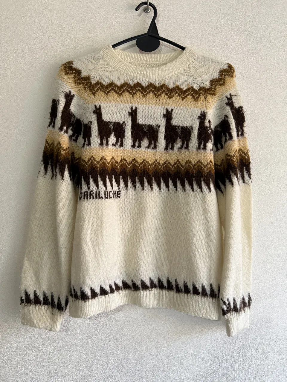 Sweater de lana tejido con cuello redondo y manga larga. Presenta un diseño tejido con figuras de llamas y patrones geométricos en tonos blanco, marrón y amarillo. En la parte delantera, tiene la inscripción "BARILOCHE" bordada.
