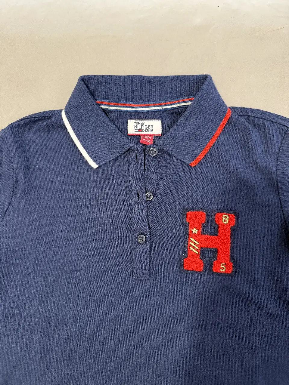 Remera polo de niño Tommy Hilfiger Denim, azul marino con detalles en rojo y blanco en el cuello. Tiene un parche con la letra H en rojo y detalles en blanco y amarillo. Ideal para un look casual y deportivo.