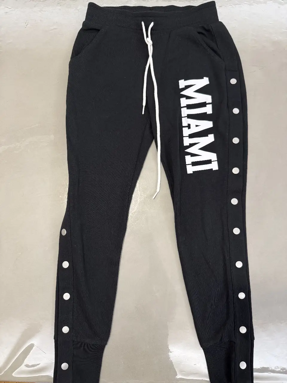 Pantalón deportivo negro de mujer, con detalle de botones en los laterales y estampado "MIAMI" en blanco. Ideal para un look casual y cómodo.