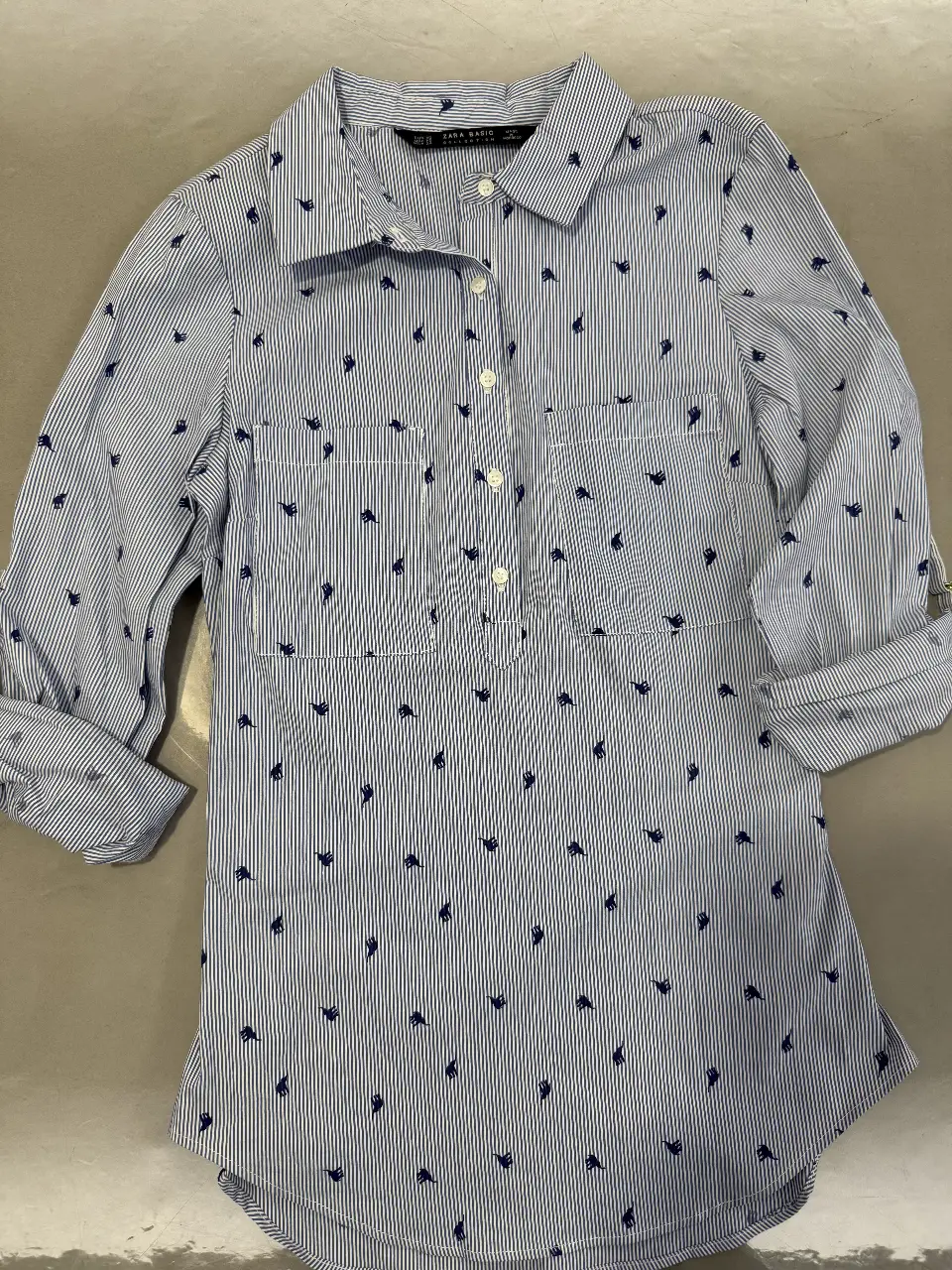Camisa de mujer a rayas celeste y blanca con estampado de pequeños dibujos azules. Tiene mangas largas con puños que se pueden doblar. Posee dos bolsillos delanteros y botones en la parte delantera. Marca Zara Basic.
