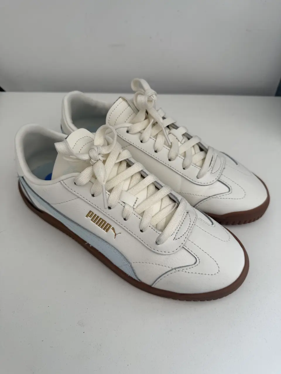 Zapatillas Puma blancas con detalles en celeste y suela marrón. Tienen el logo de la marca en dorado. Ideales para un look casual y cómodo.