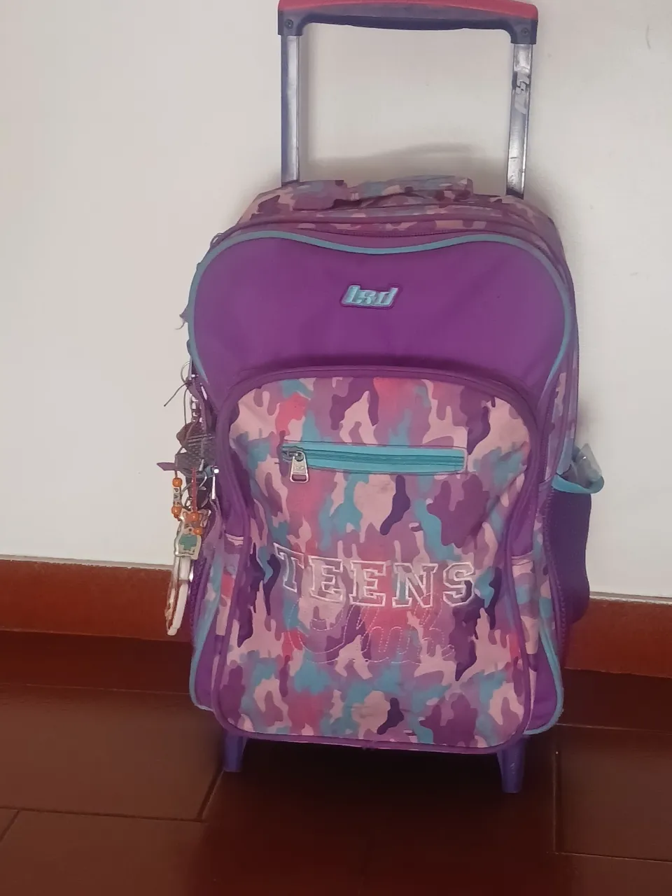 Mochila con ruedas para adolescentes, de color violeta y estampado de camuflaje. Cuenta con varios compartimentos y detalles en turquesa. Ideal para llevar libros y útiles escolares.