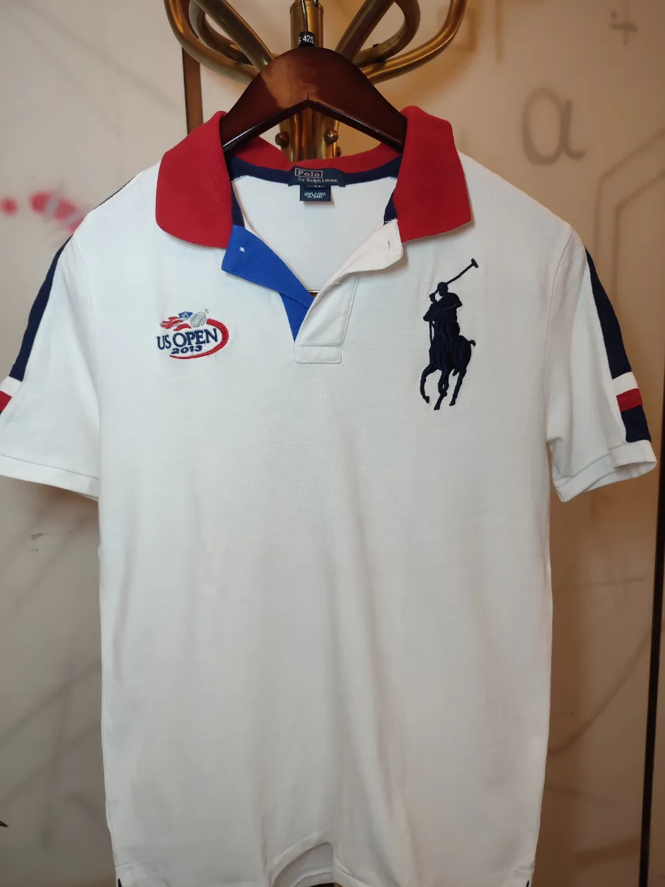 Remera blanca tipo polo con cuello y mangas rojas y azules. Logo de "US OPEN 2015" bordado en el frente izquierdo y logo de "Polo" bordado en el frente derecho. Logo de "Polo by Ralph Lauren" en la etiqueta.Chomba el talle es XL de niños para 18/20 años .Ancho de hombros 0,40 cm .Ancho de sisa  0,48 cm .Largo 0,70 cm.